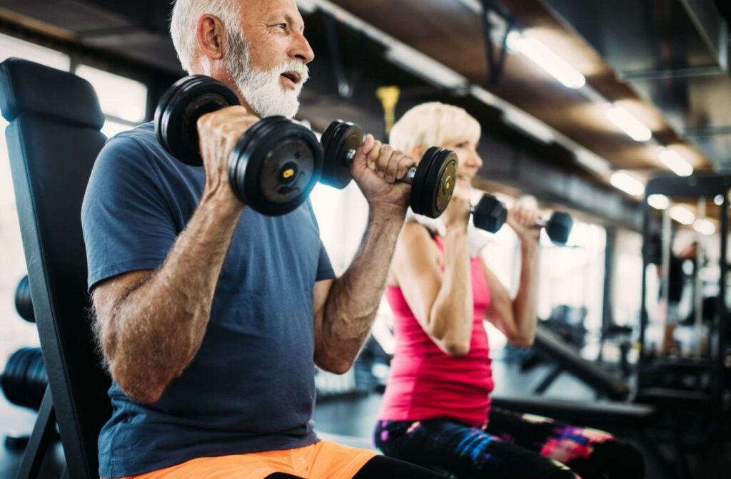 Two seniors sit side by side in a senior living community gym, doing reps with dumbbells to strengthen their muscles