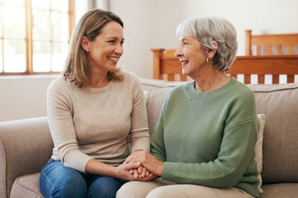 A woman and a senior sit on a couch indoors, holding hands and smiling at each other.